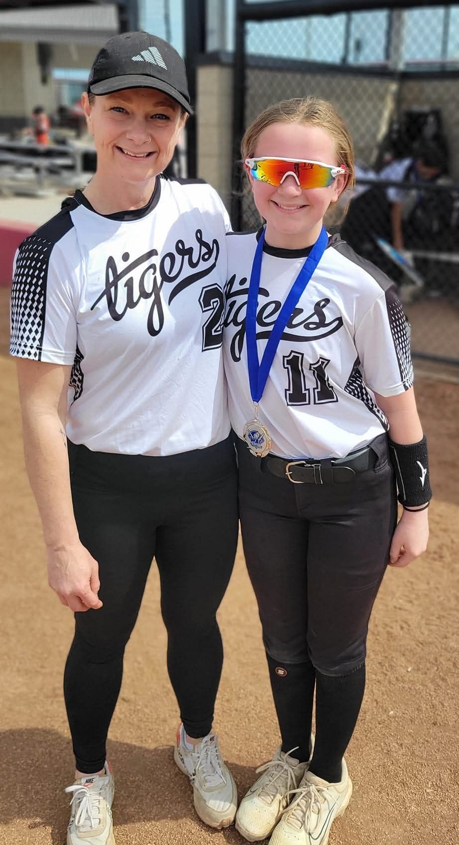 Two softball players in white jerseys and black pants pose together outdoors, smiling in team uniforms.