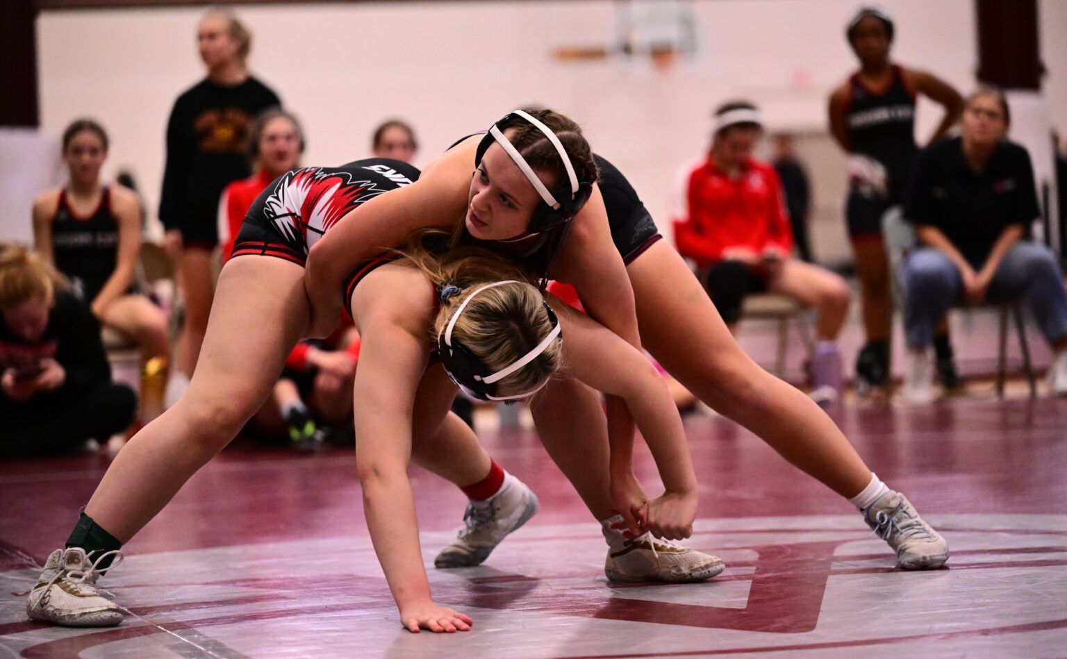 Two wrestlers crouched low on a mat, locked in a grapple during a match with spectators in the background