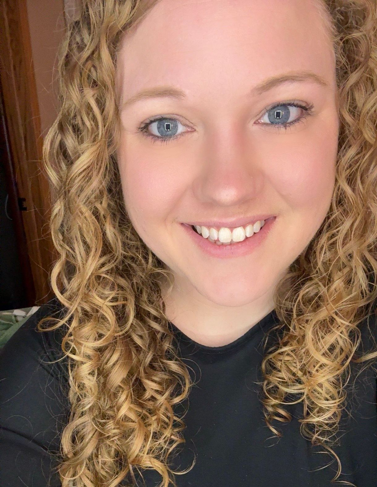 Smiling person with curly blonde hair wearing a black top against a dark background