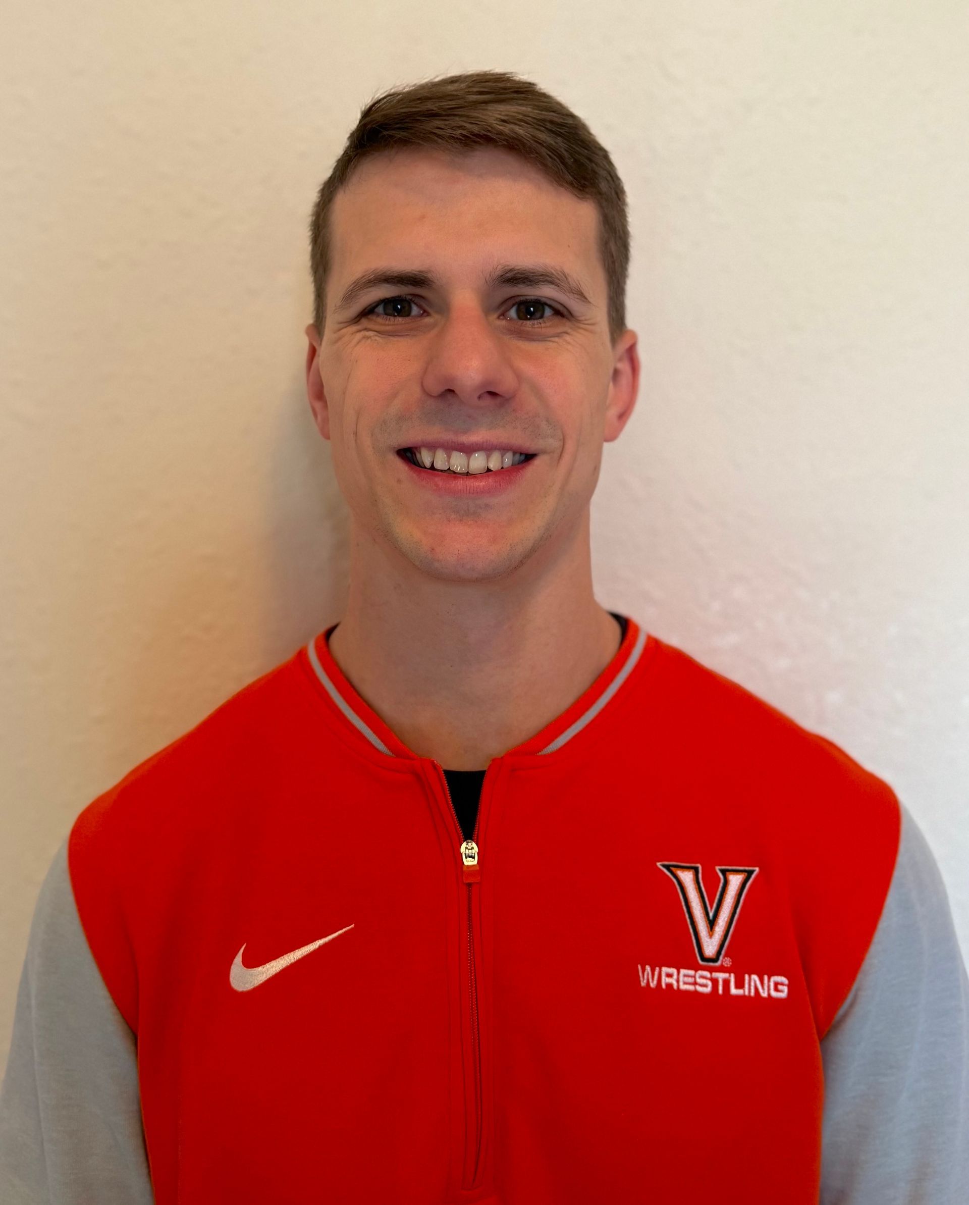 Smiling person in a red and gray wrestling jacket against a plain wall