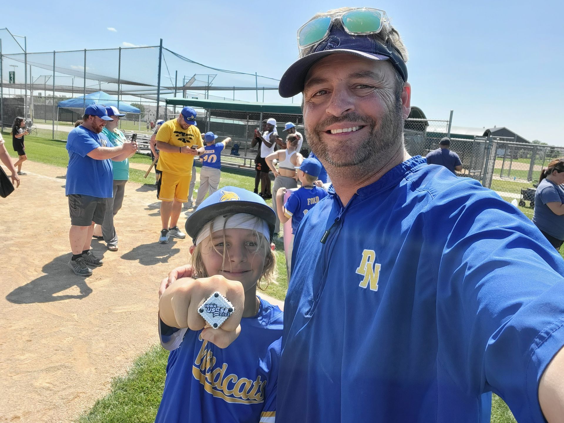 Two baseball fans in blue gear smiling at a sunny ballfield selfie