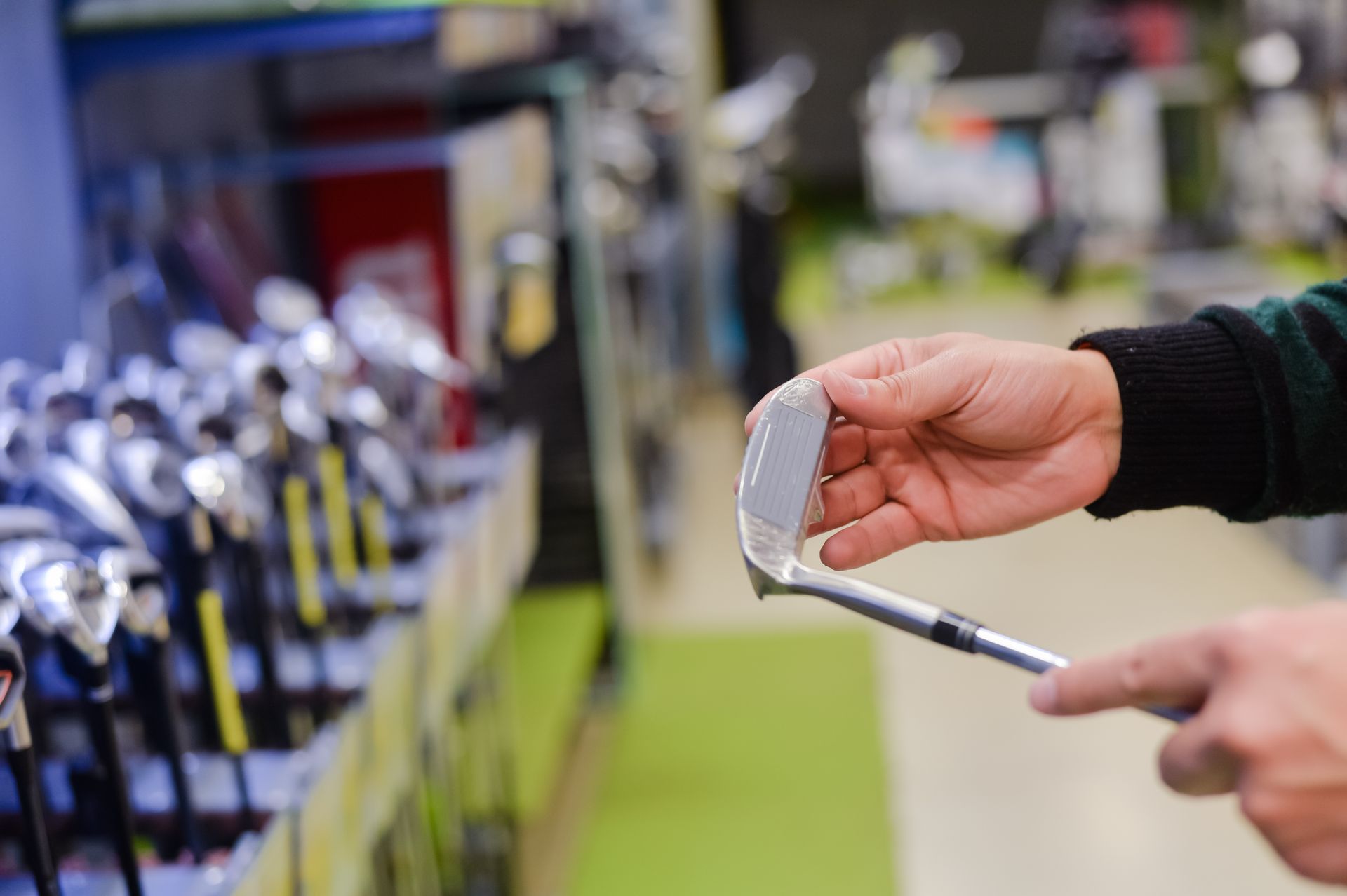 Hands inspecting a golf iron head with rows of clubs displayed behind in a golf shop