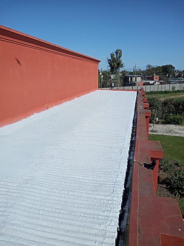 Techo plano revestido de blanco junto a un alto muro rojo, con una zona de césped visible a la derecha bajo un cielo azul.