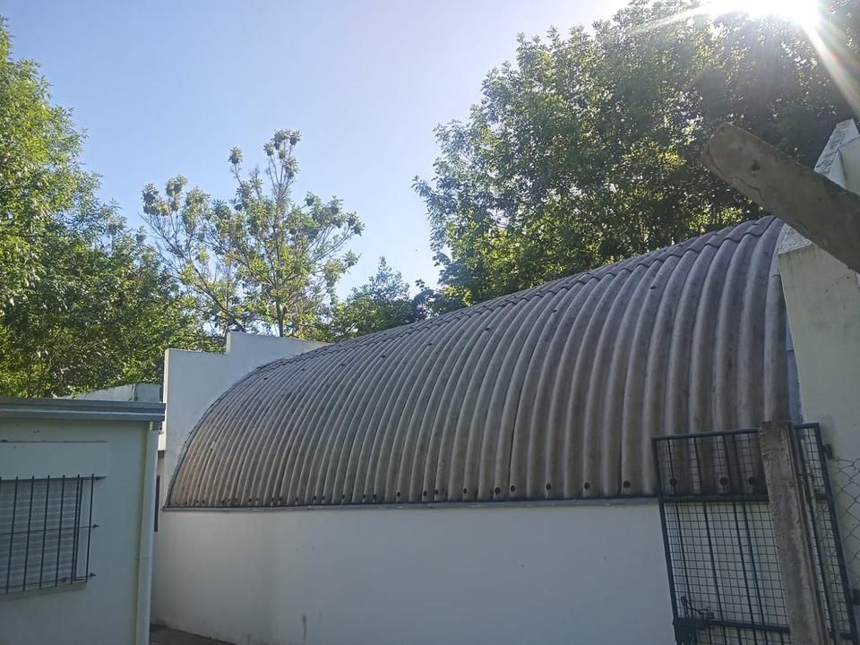 Edificio blanco con techo de metal corrugado, frente a árboles y cielo azul.