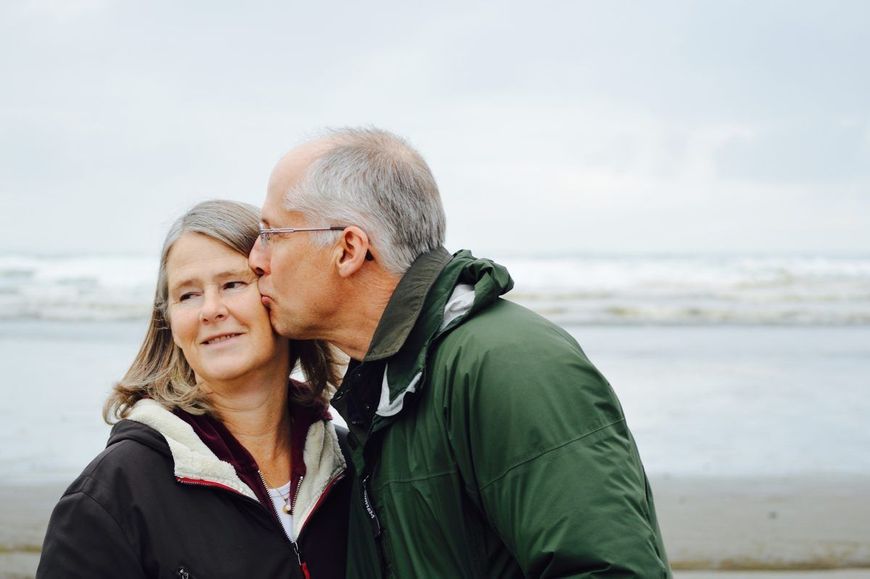 Two people on a windy beach, one kissing the other on the cheek, with the ocean behind them