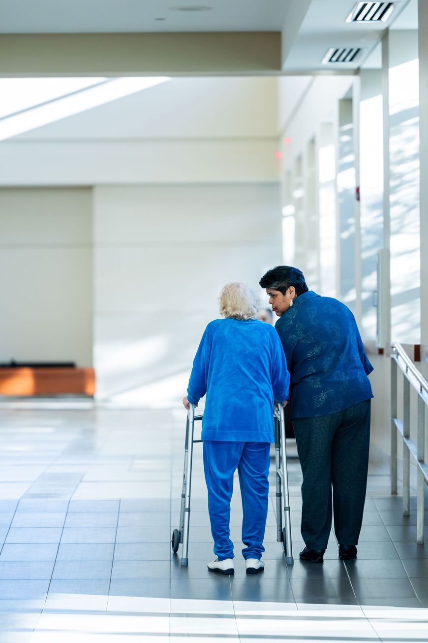Two people walking down a hospital corridor, one using a walker