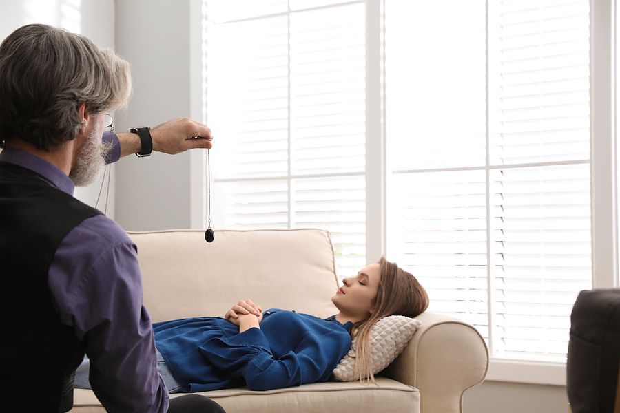 Man holding a pendulum hypnotizes a person lying on a couch near a window.