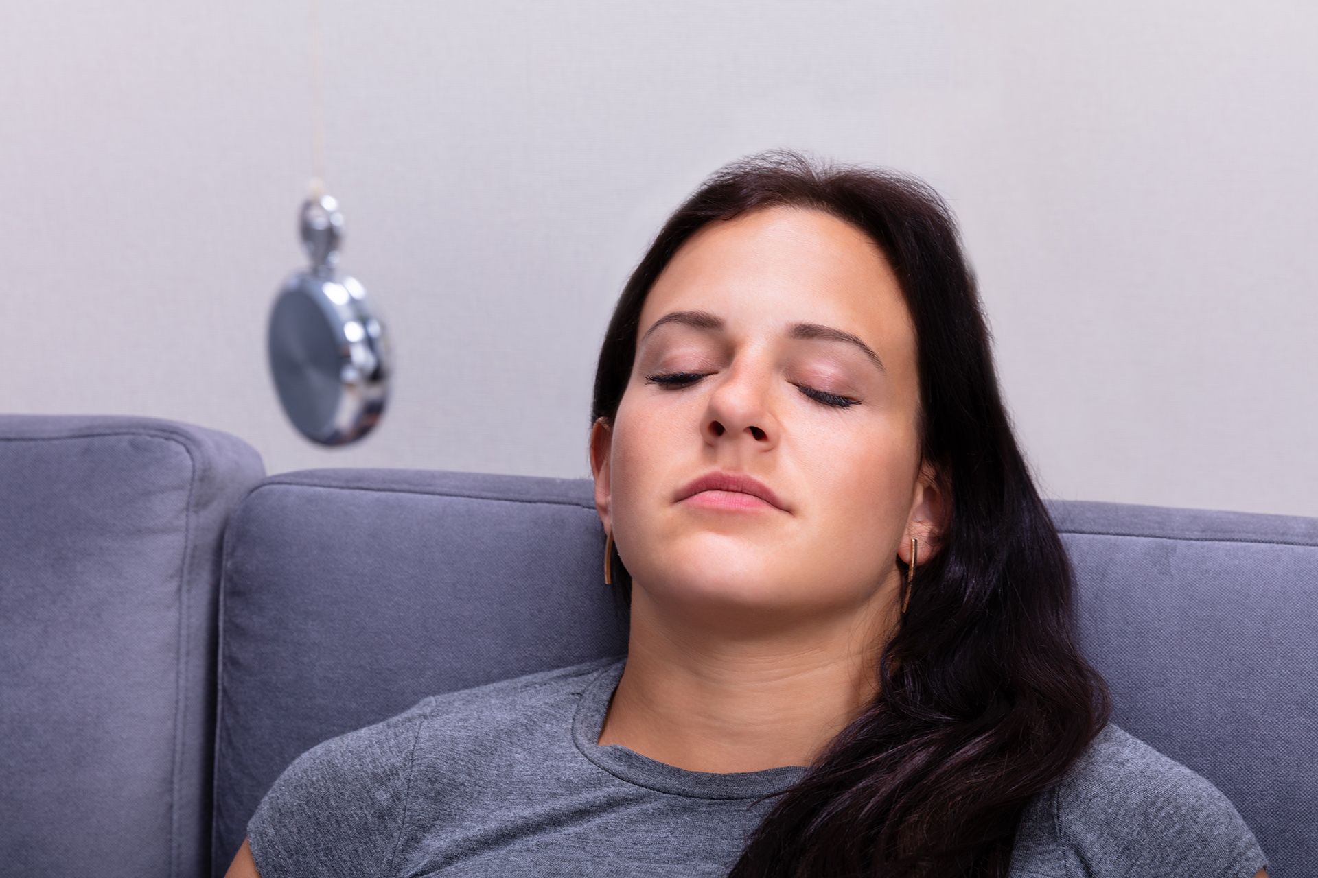 Woman on a couch with eyes closed, seemingly hypnotized by a swinging pocket watch. Gray setting.