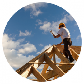 A construction worker in a hard hat hammers a wooden roof truss against a blue, cloudy sky.
