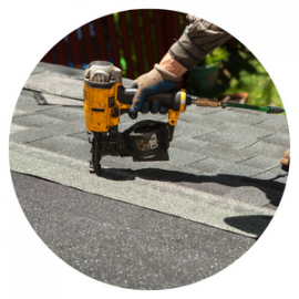 A worker uses a yellow pneumatic nail gun to install asphalt shingles on a roof.