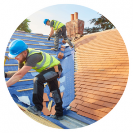 Two construction workers wearing safety gear install blue underlayment and tiles on a residential roof.