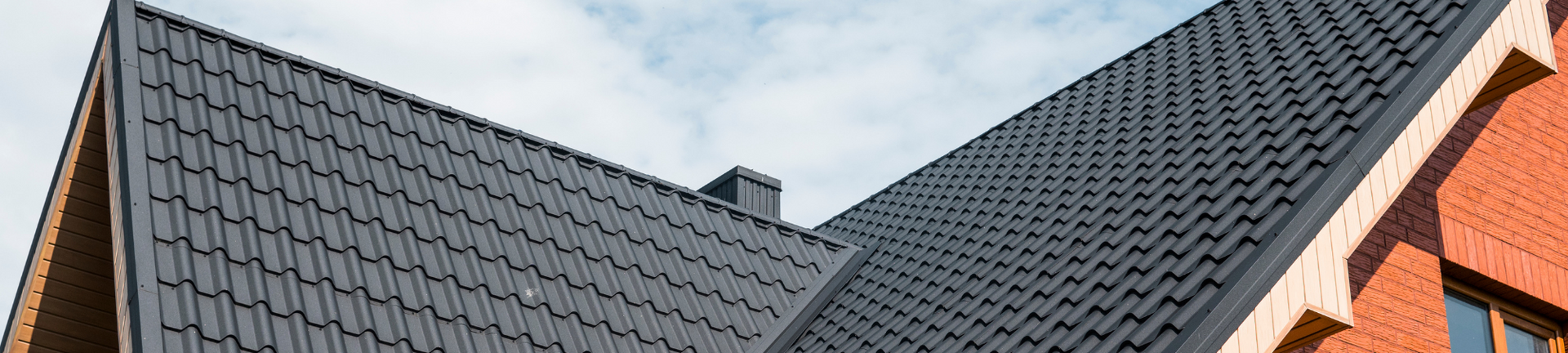 Close-up of a house roof with dark gray metal tiles, a small chimney, and brick walls under a cloudy sky.