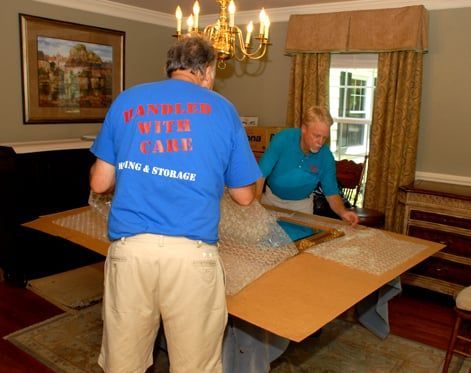 Two movers wrapping a picture frame in bubble wrap on a table inside a dining room.