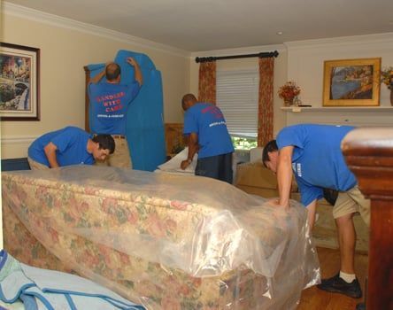 Movers wrapping a mattress in plastic in a living room, blue shirts.