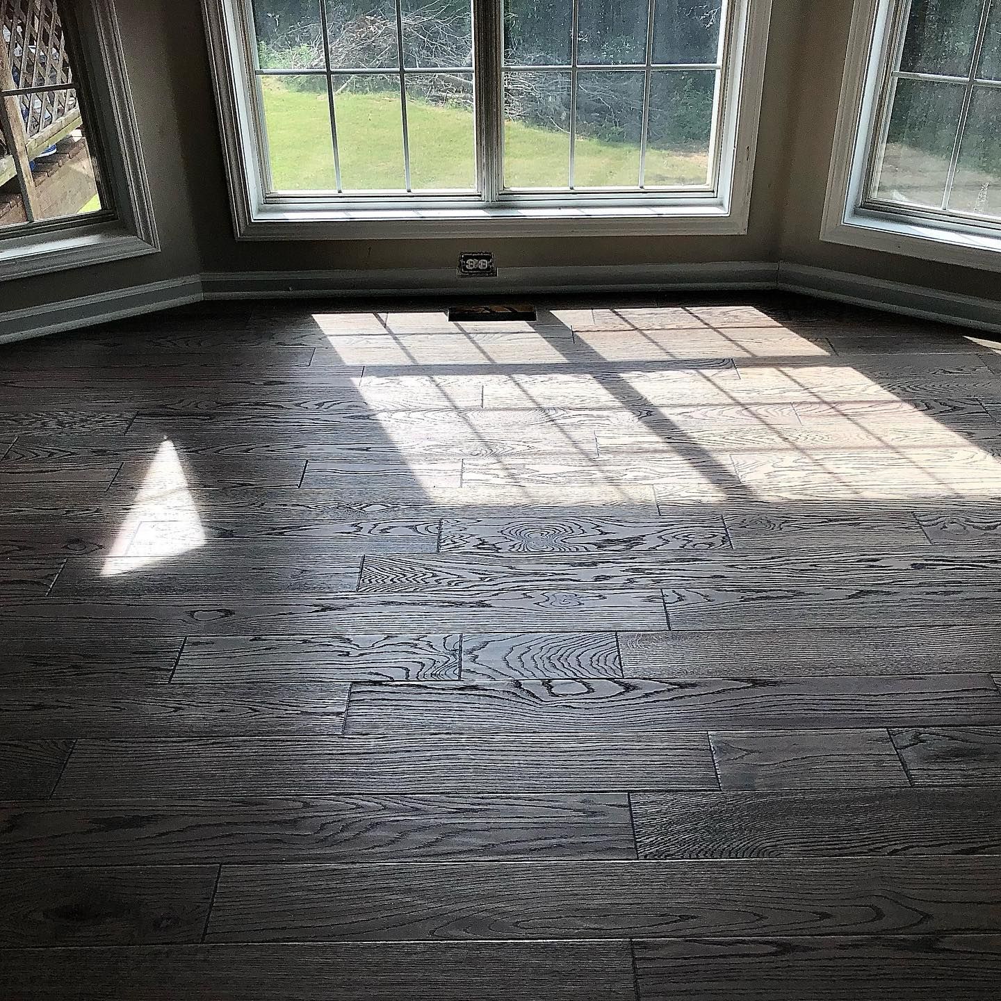 Sunlit empty room with dark hardwood floors and bay windows overlooking a green yard