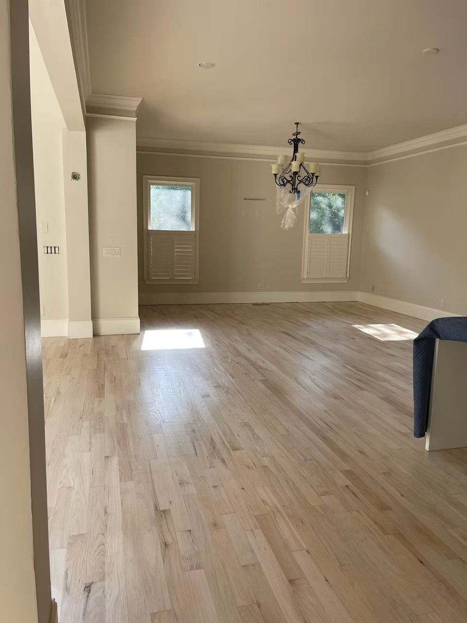 Empty living room with light wood floors, cream walls, windows, and a chandelier.