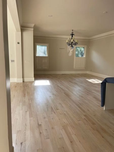 Empty living room with light wood floors, cream walls, windows, and a chandelier.