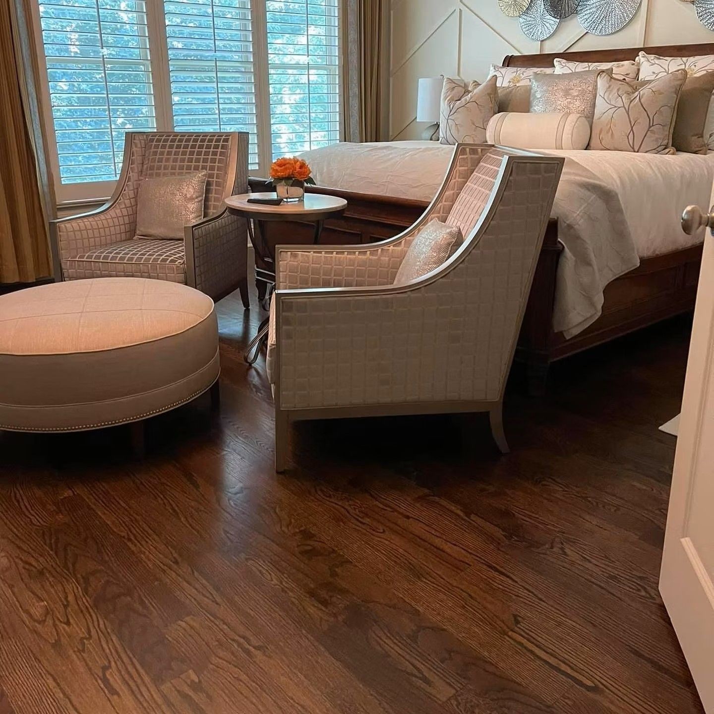 Elegant bedroom with wicker chairs, round ottoman, and a bed by a window with blue blinds.