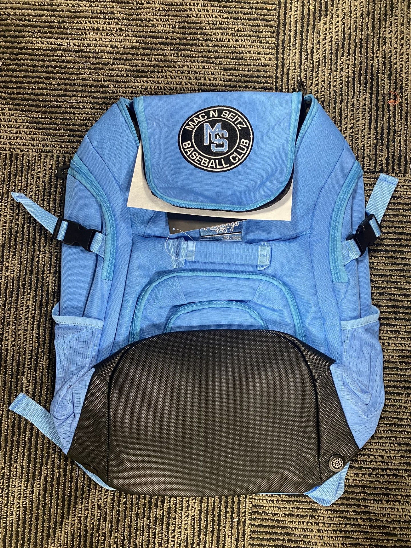 A light blue backpack sits on a brown, textured carpet. It features a circular