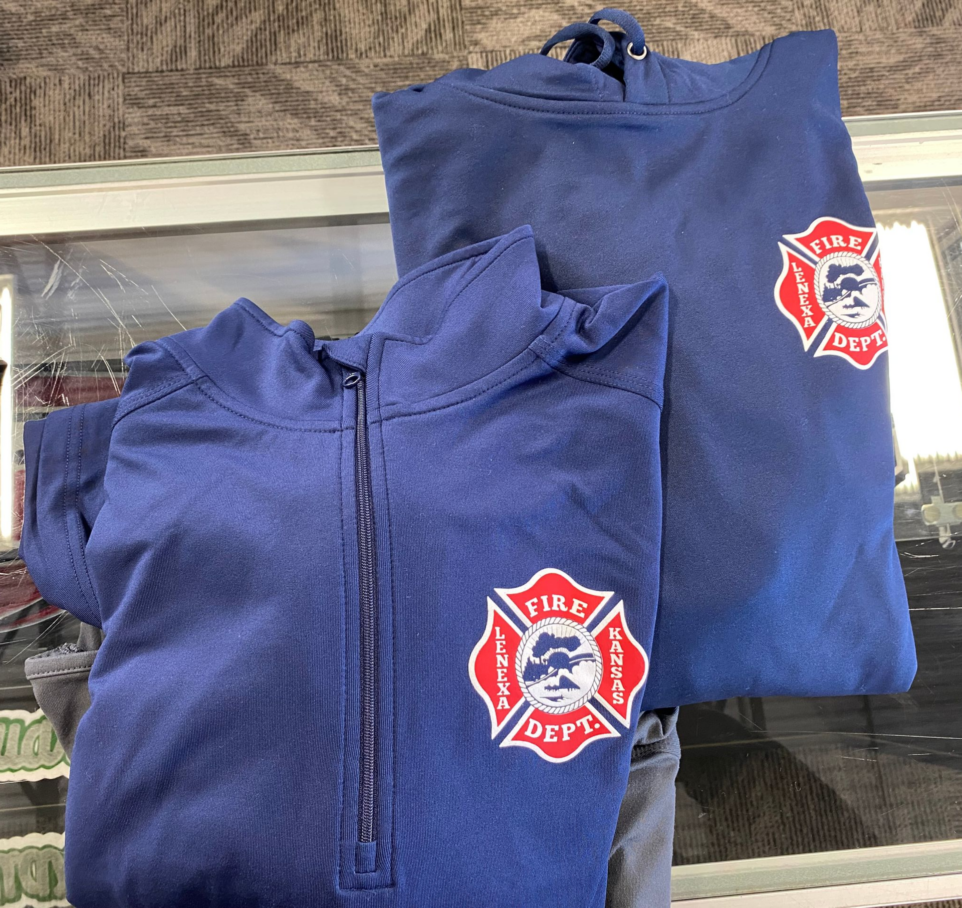 Blue firefighter zip-up jacket and hoodie with fire department logo, displayed on a glass surface.