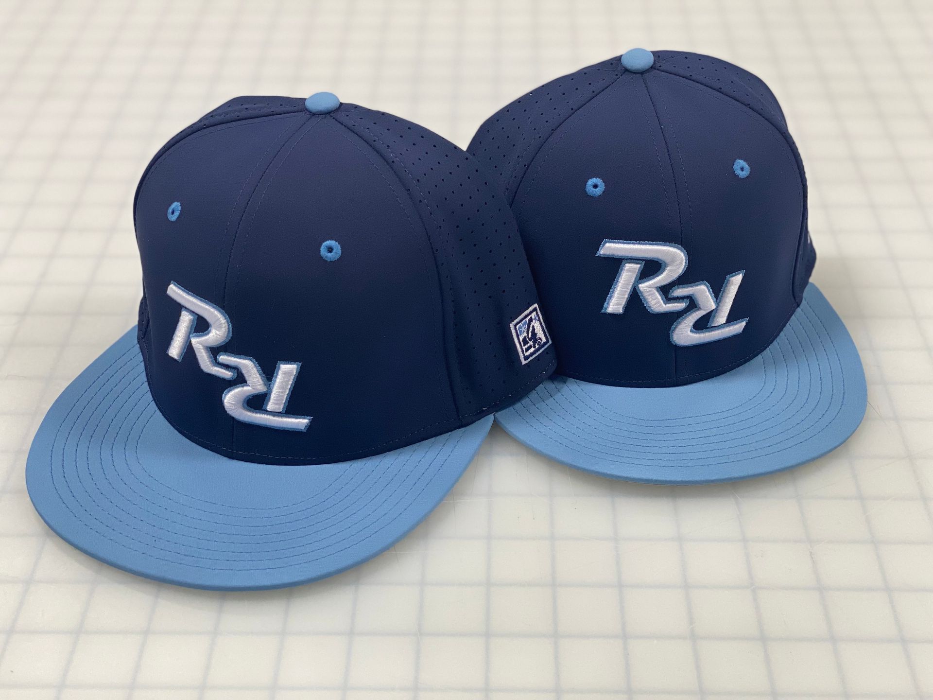 Two navy blue baseball caps with light blue brims, featuring the letters