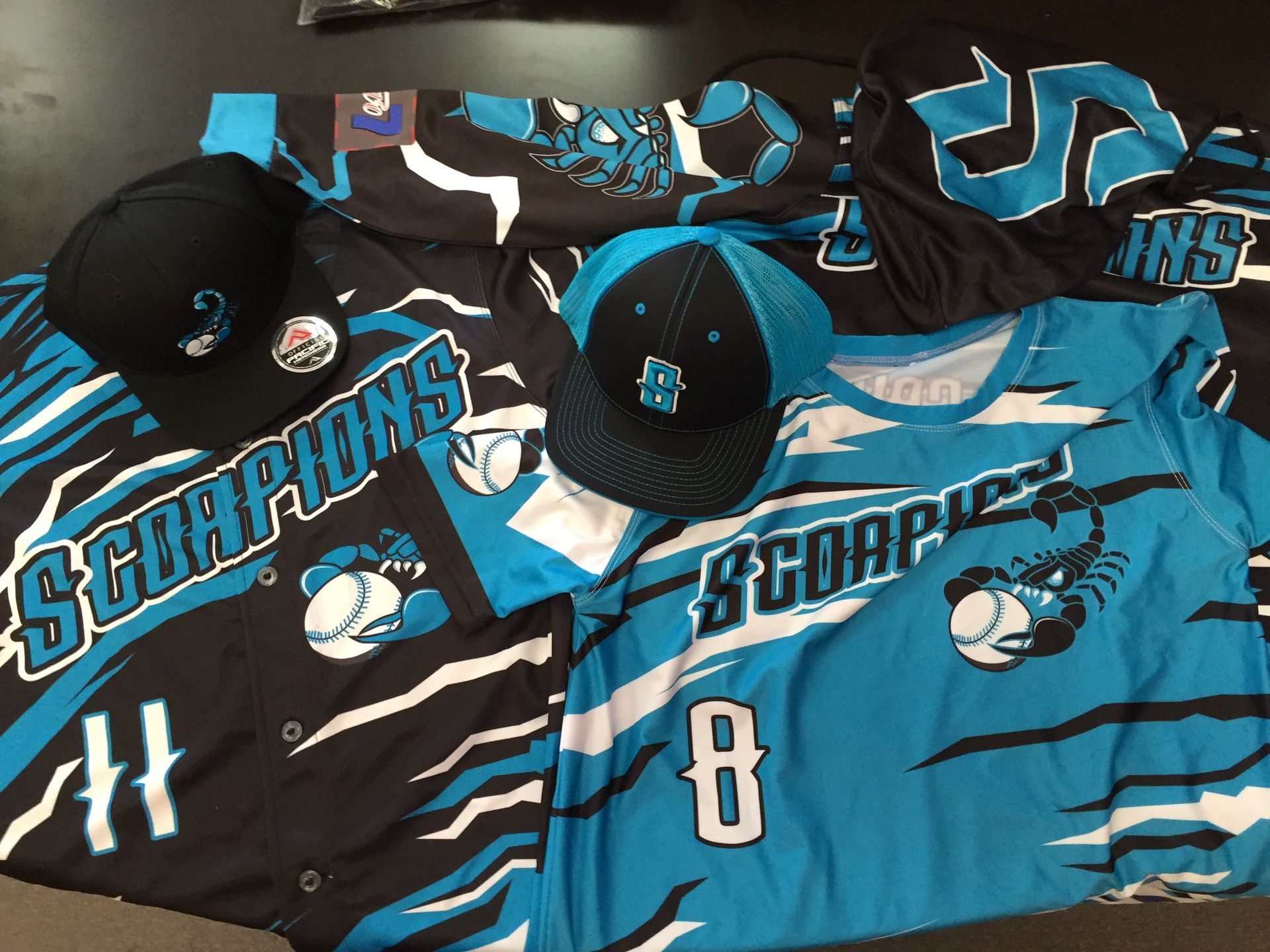 Baseball team jerseys and hats. Teal and black Scorpions team gear on a dark surface.