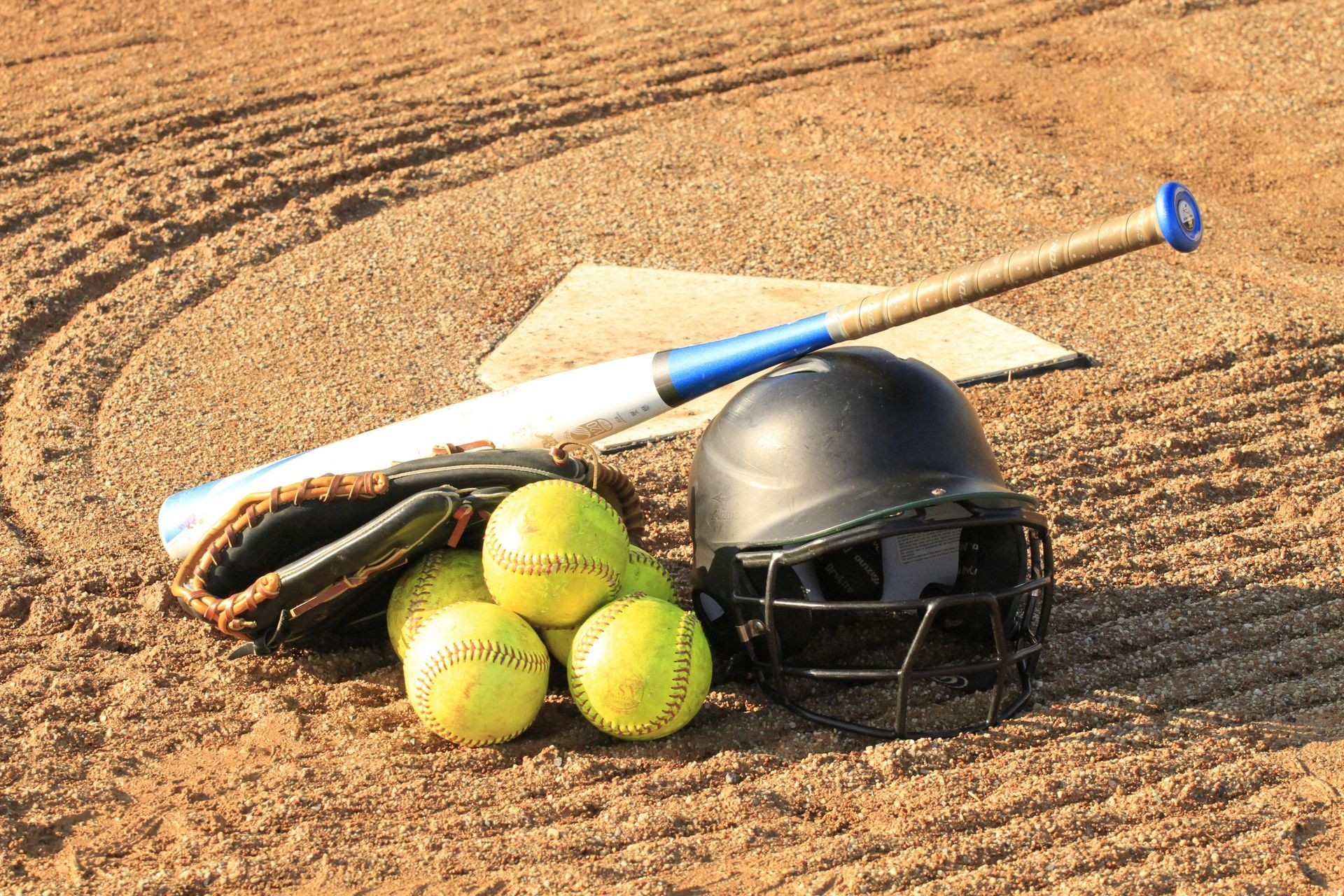 Softball equipment on a dirt field: balls, bat, helmet, and glove.