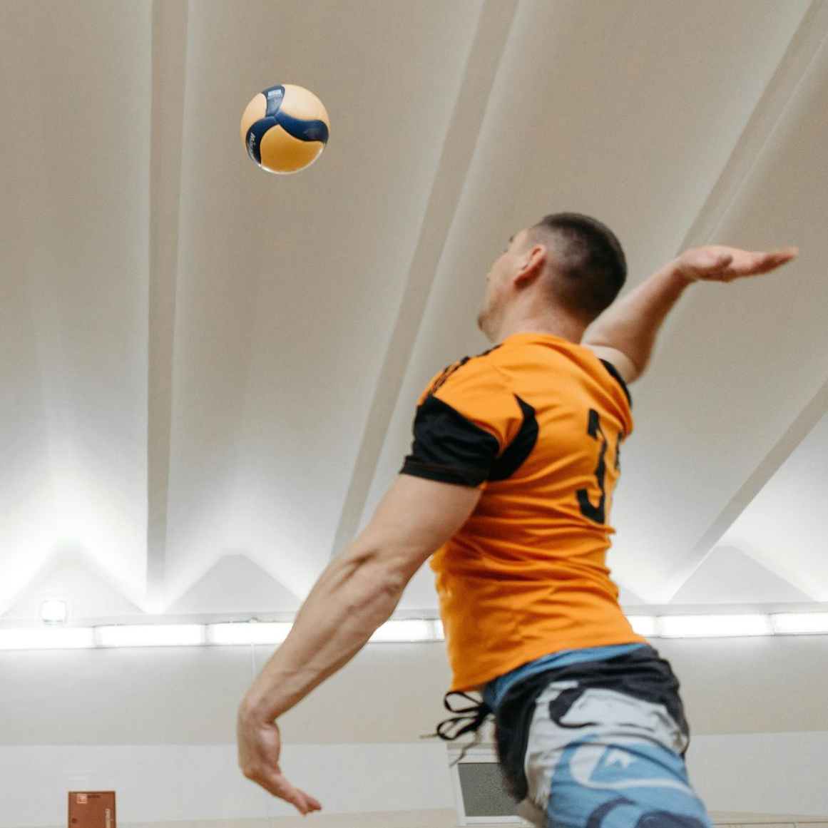 Man in orange shirt tossing a ball upwards. Indoor setting, bright lights, arm extended.