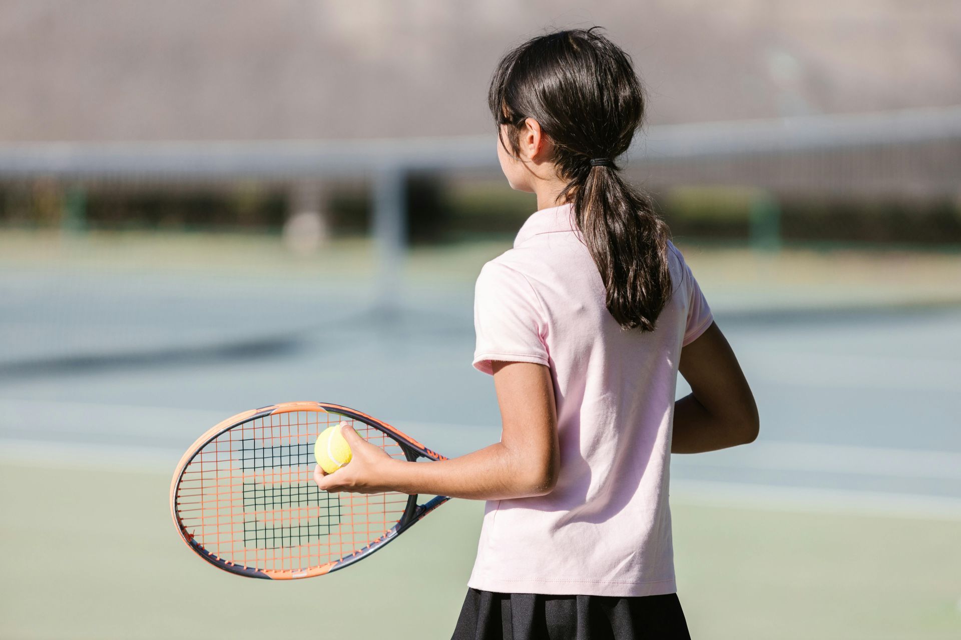 Girl holding tennis ball and racket on a tennis court, ready to serve.