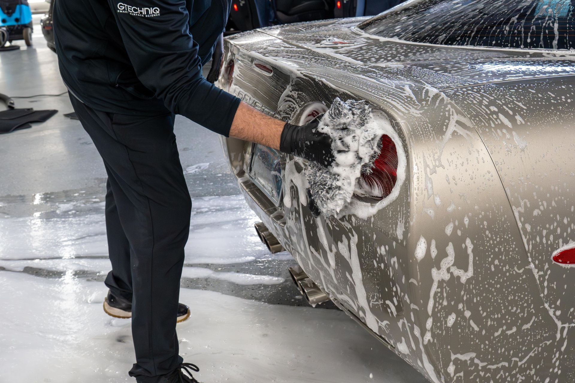 Person wearing gloves washing a silver sports car with soapy sponge.