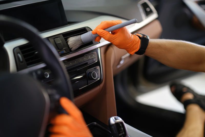 A person cleaning a car's dashboard with a brush and wearing orange gloves.
