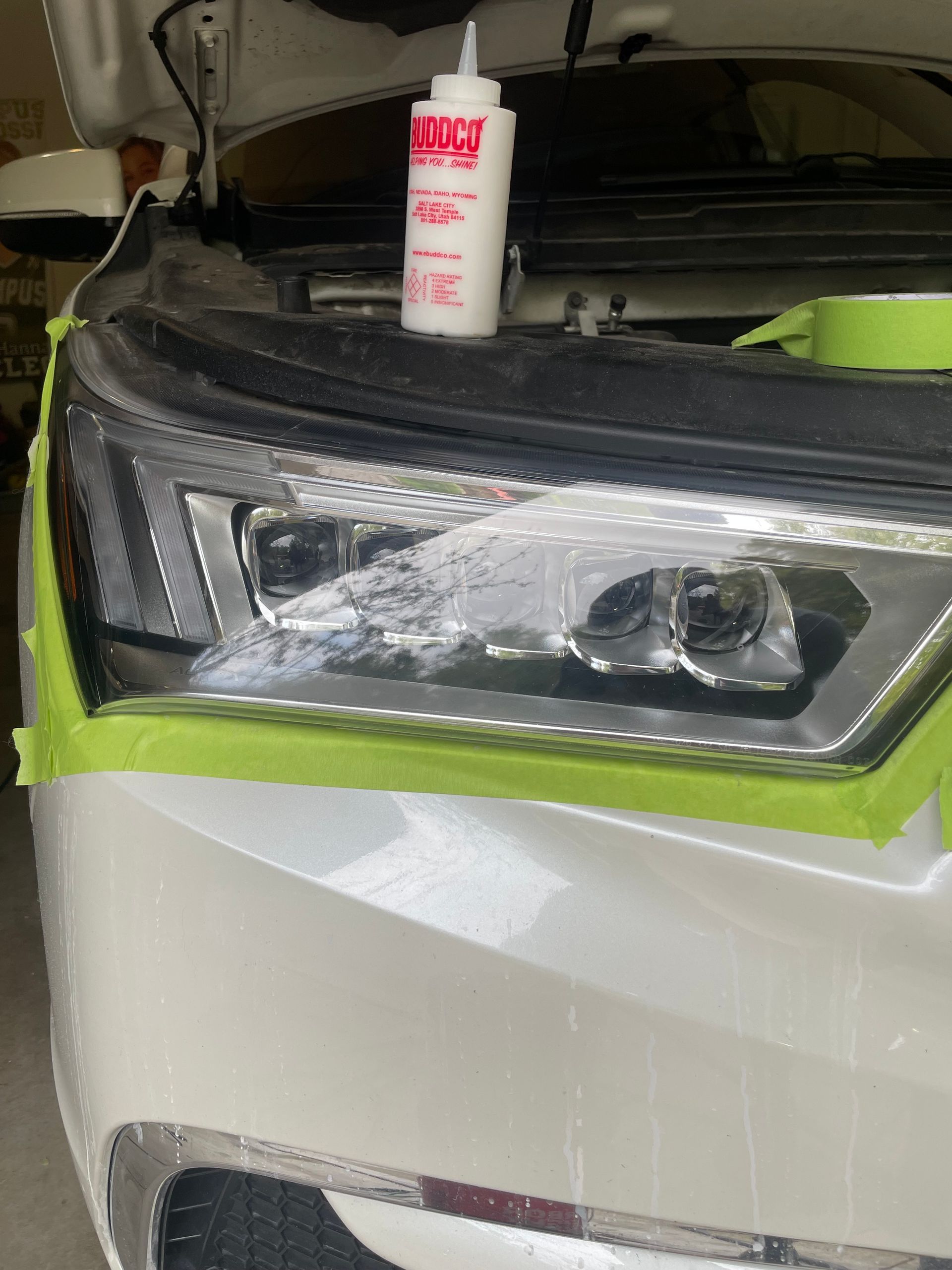 Headlight being restored with polish; car hood open, taped, and white. Bottle of polish visible.