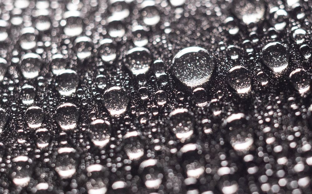 Water droplets on a dark, textured surface, reflecting light and creating a glistening pattern.