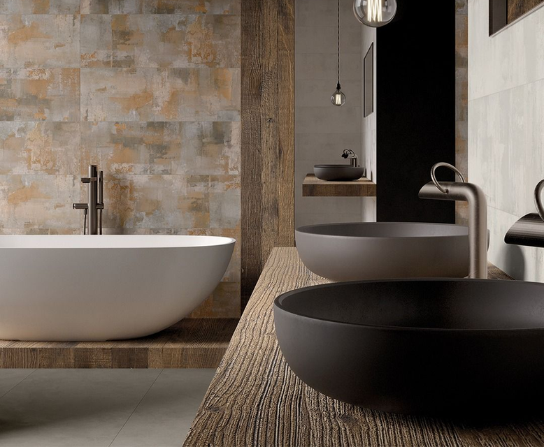 Bagno con elementi rustici in legno e accessori moderni: vasca bianca, lavandini scuri e pareti piastrellate con effetto anticato.