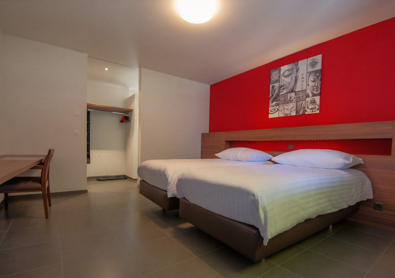 Chambre d'hôtel moderne avec deux lits blancs, un mur d'accent rouge, un bureau et une porte ouverte.