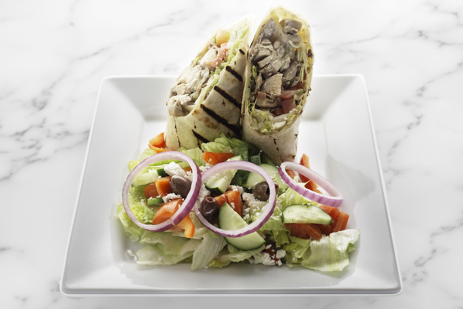 A white plate holds two halves of a grilled chicken wrap and a side salad with lettuce, tomatoes, olives, and red onion.