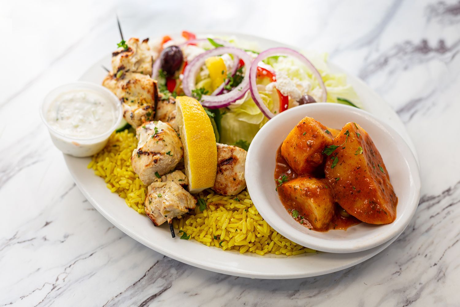 A Greek-style chicken souvlaki platter with yellow rice, salad, a lemon wedge, tzatziki, and stewed potatoes in a bowl.