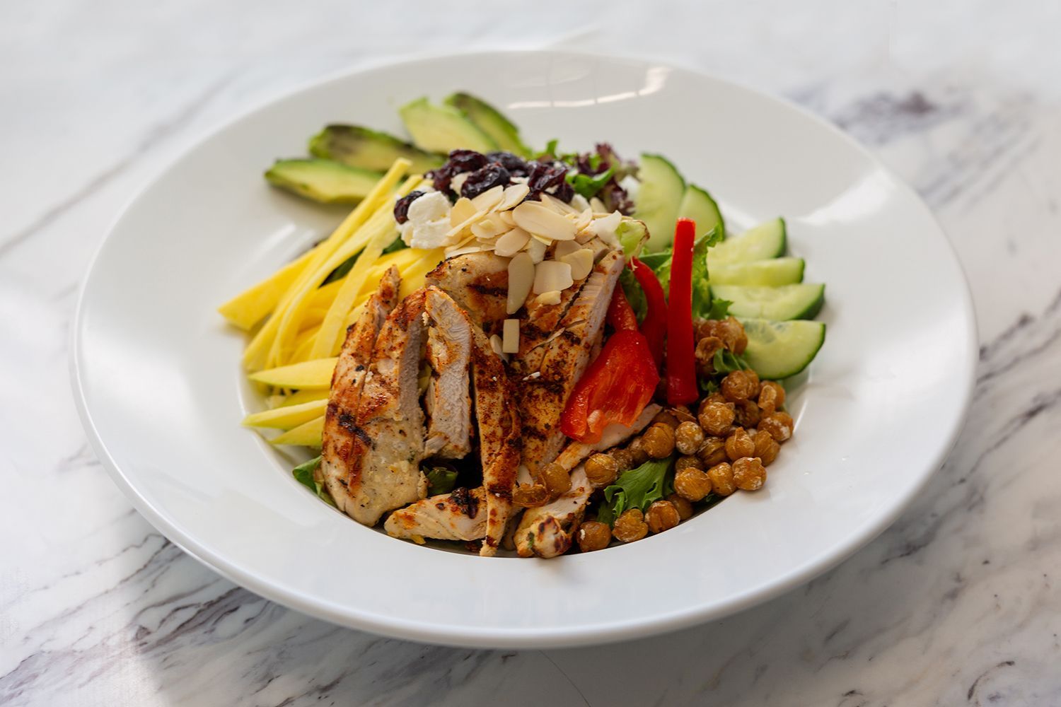 A salad in a white bowl featuring grilled chicken, sliced mango, avocado, cucumber, chickpeas, and topped with nuts.