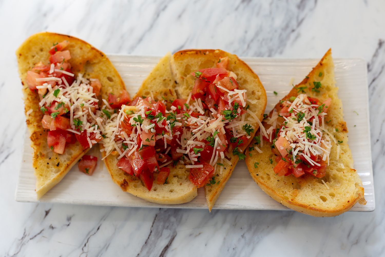 Three pieces of toasted garlic bread topped with chopped fresh tomatoes, shredded parmesan cheese, and herbs on a tray.