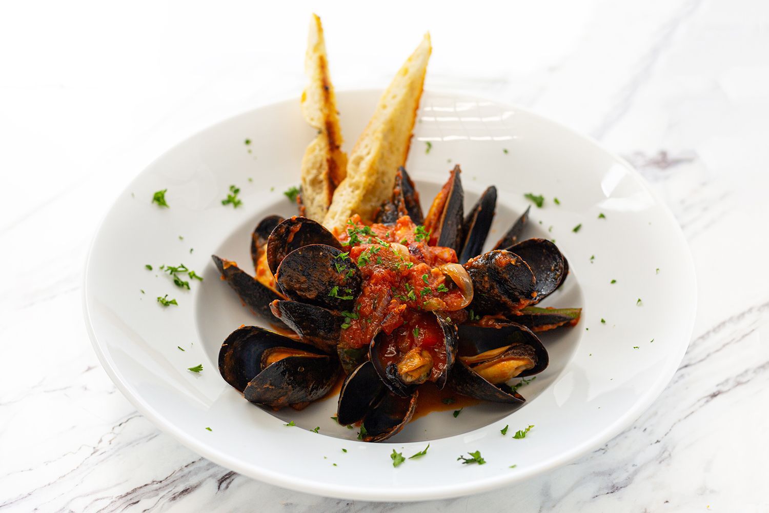 A white bowl containing mussels in a tomato sauce, garnished with herbs and served with two pieces of toasted bread.