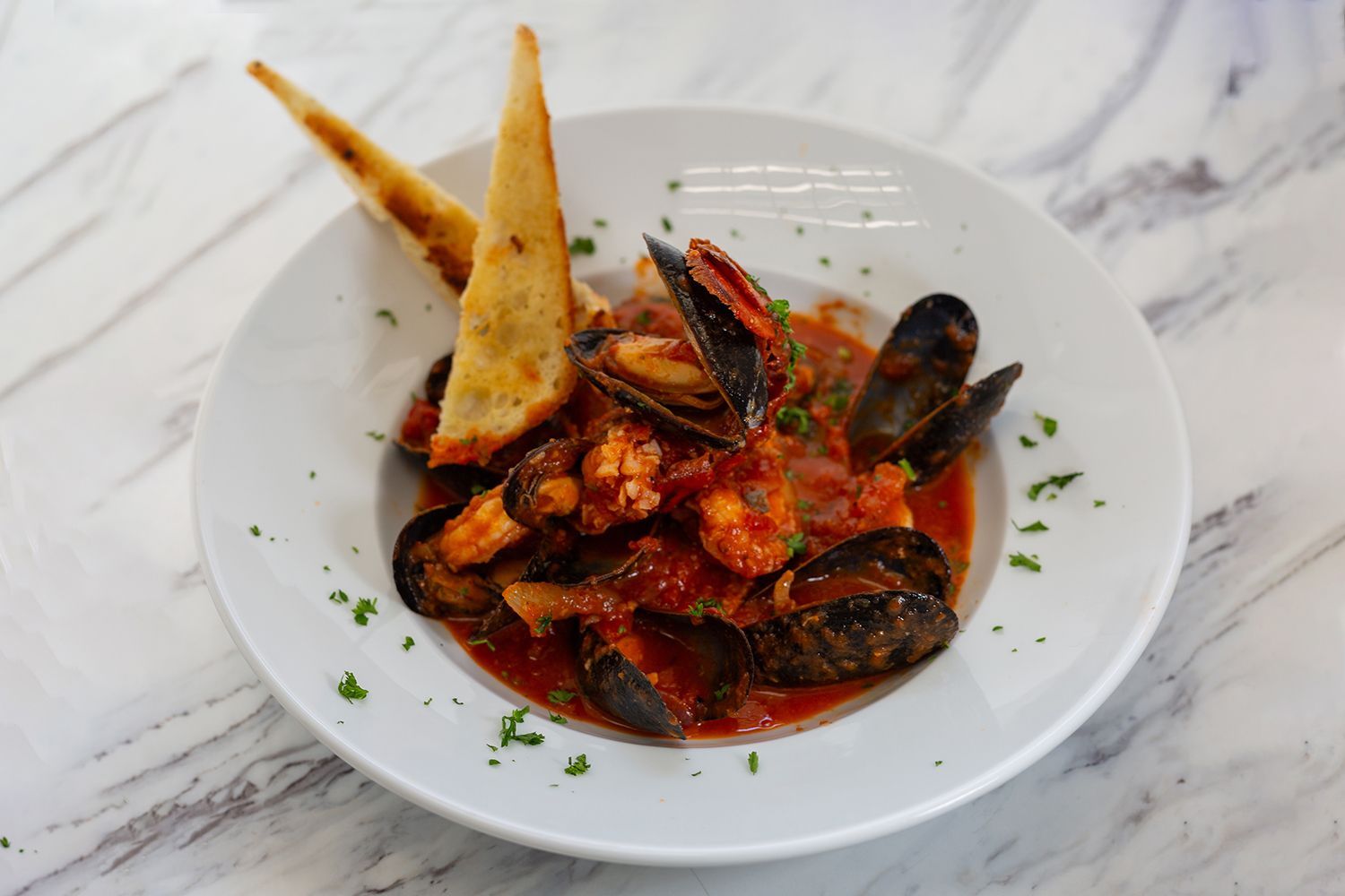 A bowl of seafood stew with mussels and shrimp in a red tomato sauce, served with two toasted bread points.
