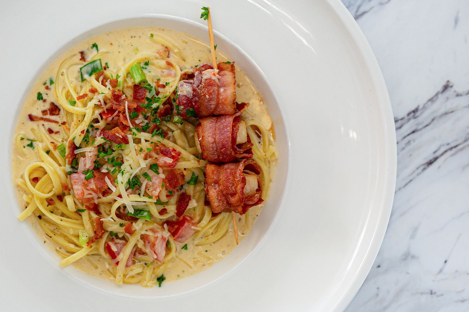 A bowl of creamy pasta topped with bacon bits, herbs, and a skewer of three bacon-wrapped scallops on a marble table.