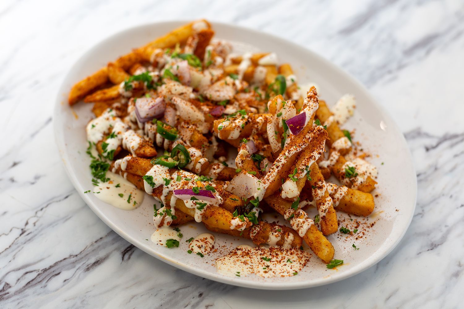 A plate of seasoned french fries drizzled with white sauce, topped with red onion, jalapeños, and fresh herbs.