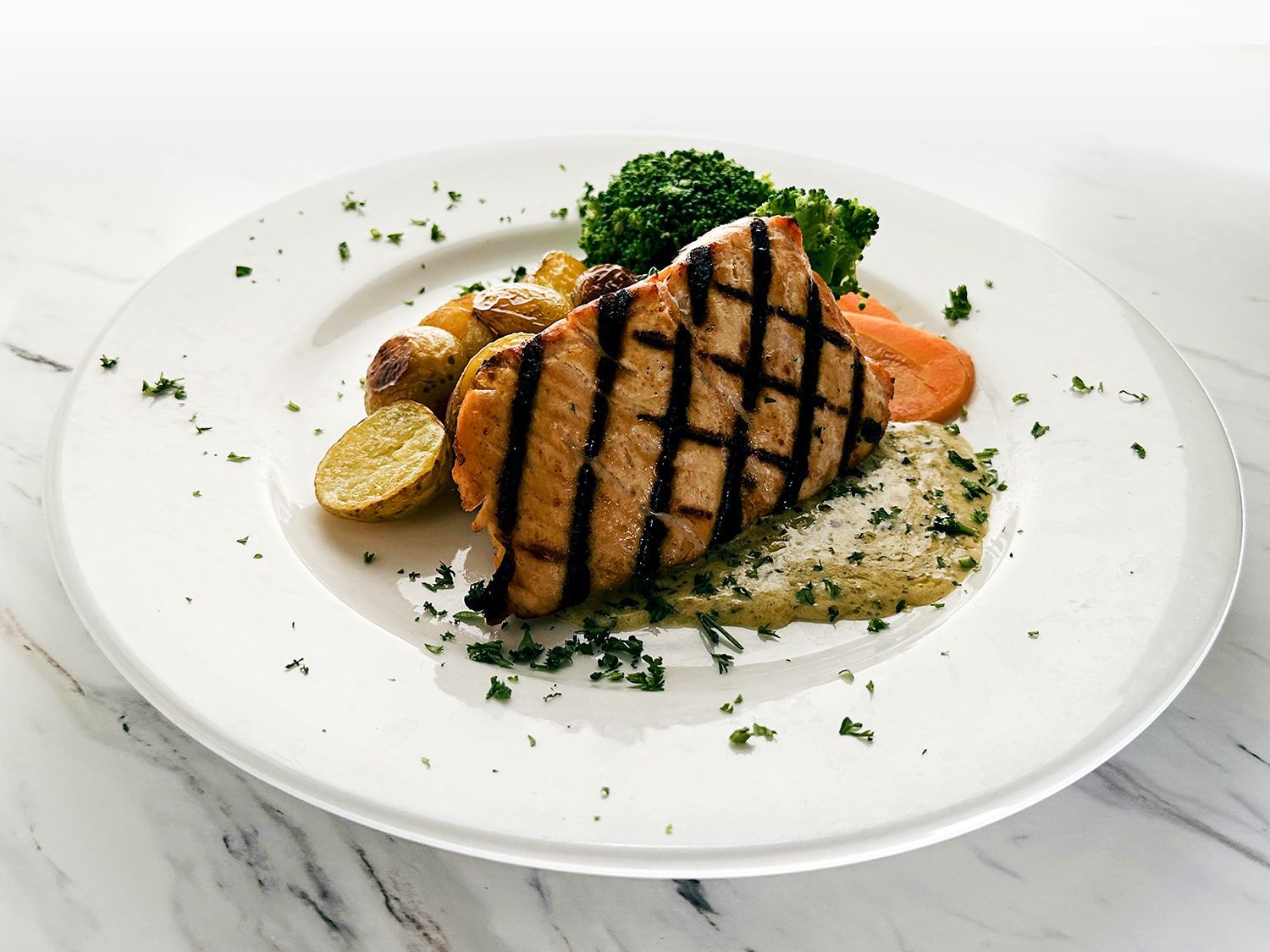 A plated grilled pork chop served with roasted potatoes, steamed broccoli, carrots, and a side of pureed greens.