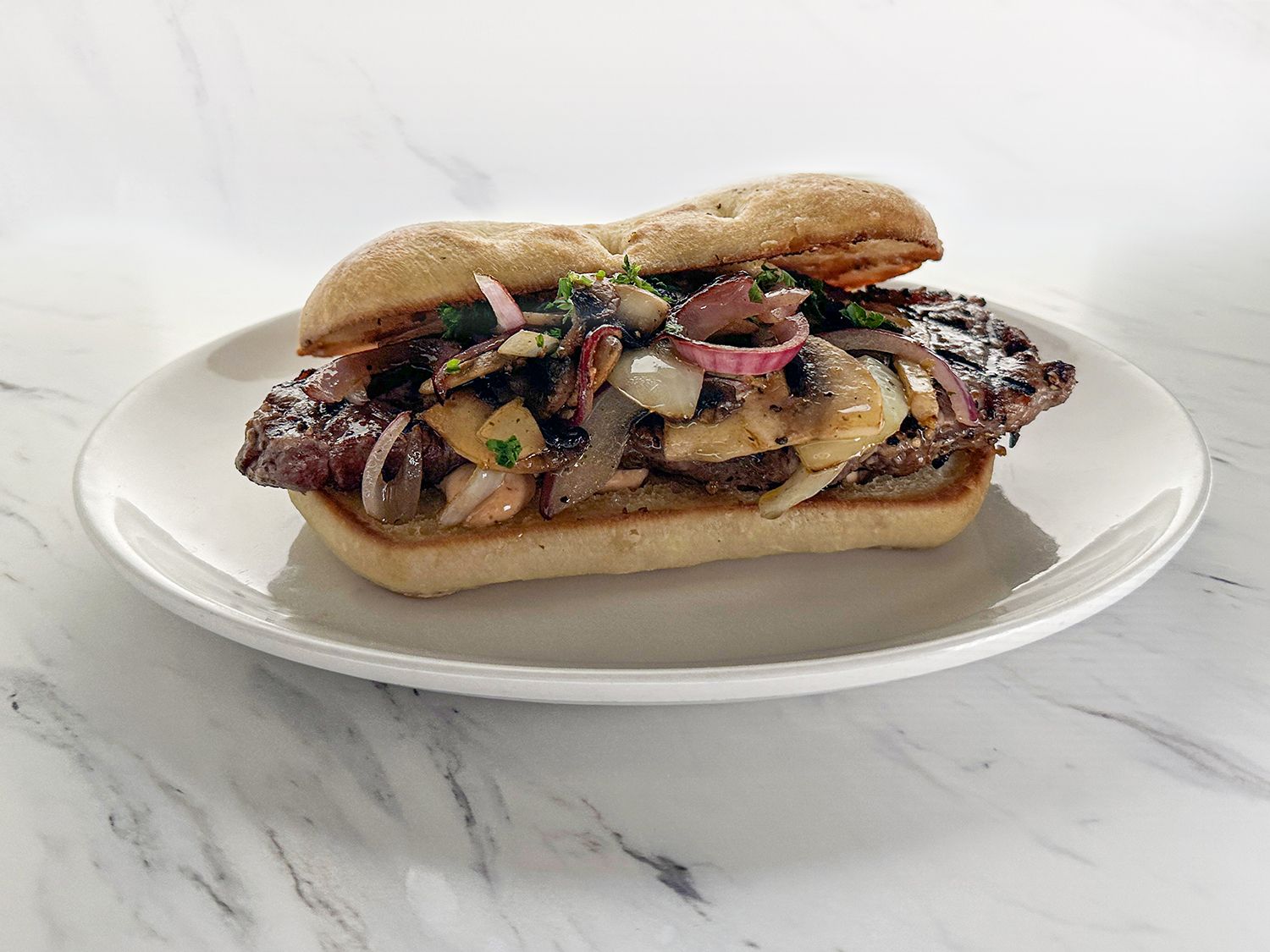 A steak sandwich with grilled onions and cheese on a toasted bun, served on a white plate against a marble background.