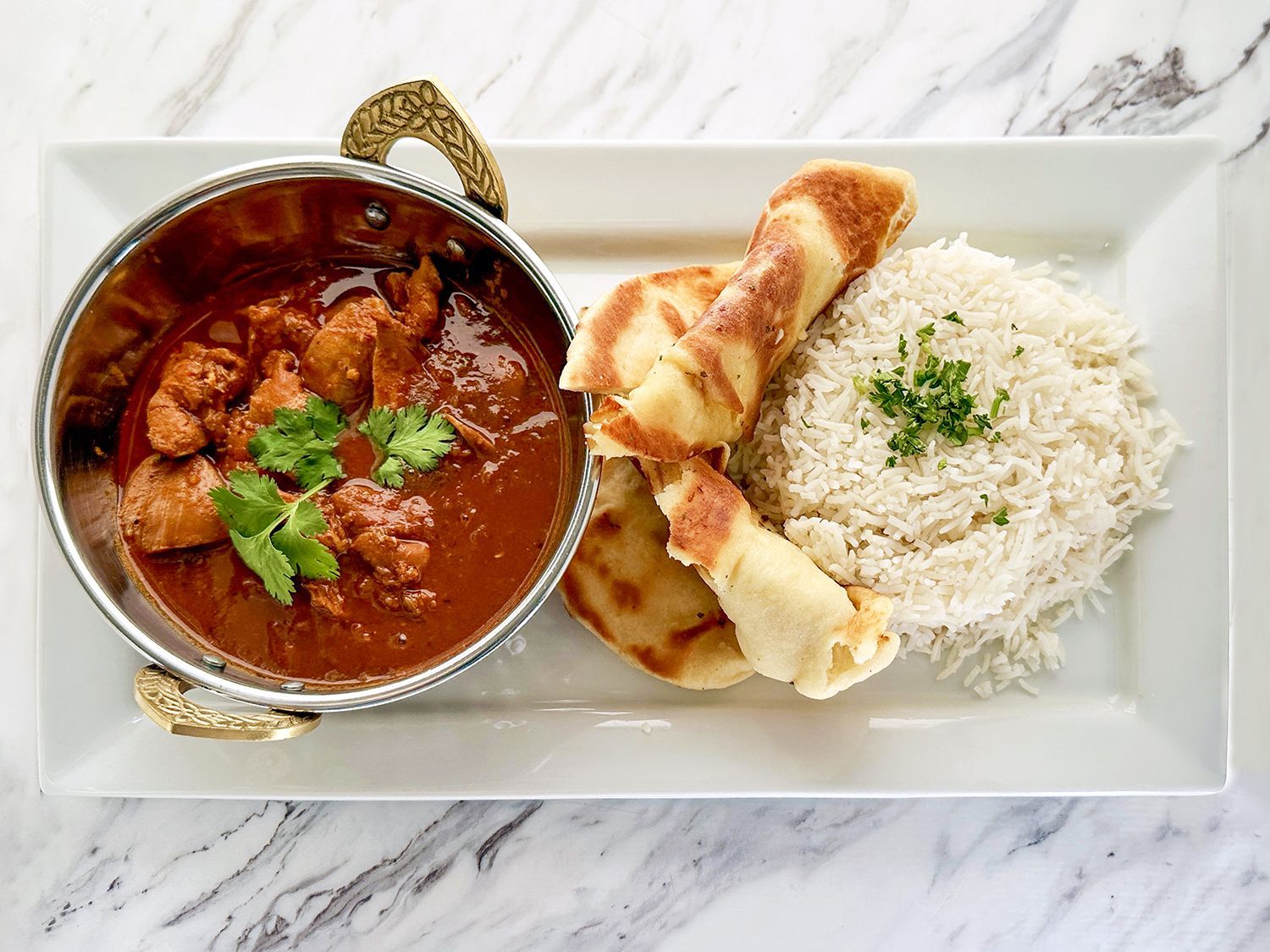 A bowl of chicken curry garnished with cilantro, served with flatbread and white rice on a rectangular plate.