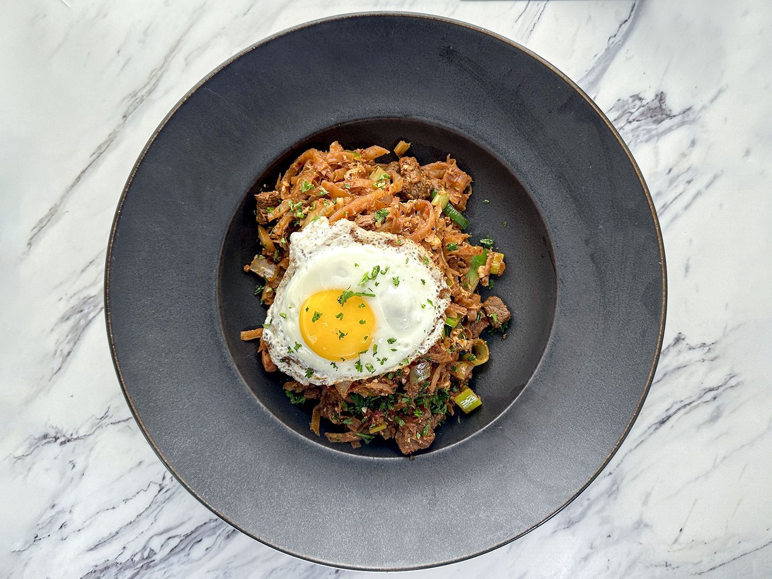 A dark textured bowl on a marble surface containing stir-fried food topped with a sunny-side up egg and herbs.