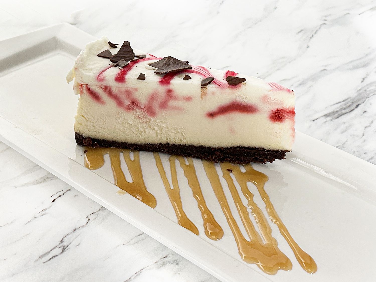 A slice of cheesecake with red swirls, chocolate shavings, and caramel drizzle on a white rectangular plate.