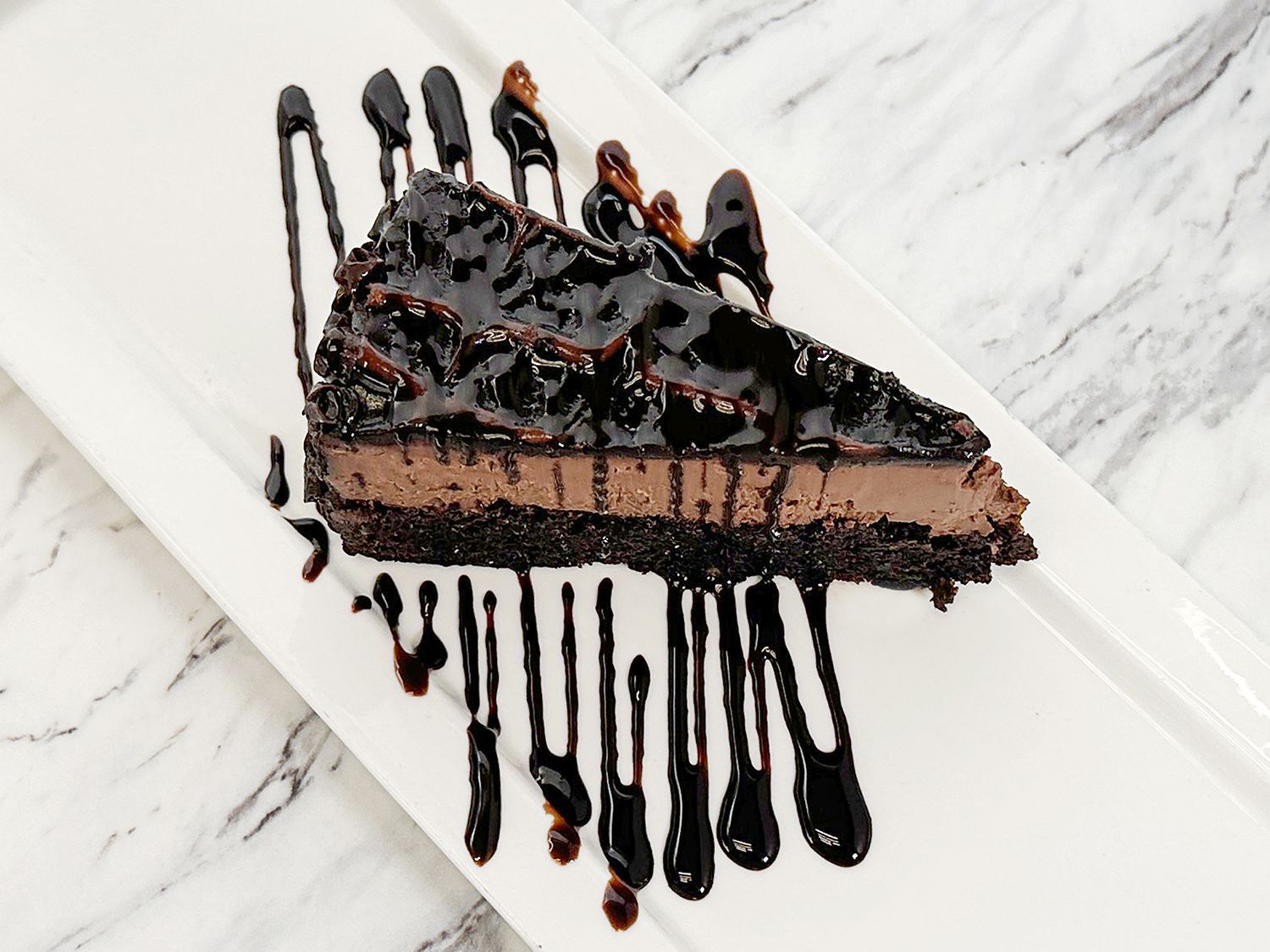 A slice of chocolate cheesecake drizzled with chocolate sauce on a white rectangular plate against a marble background.