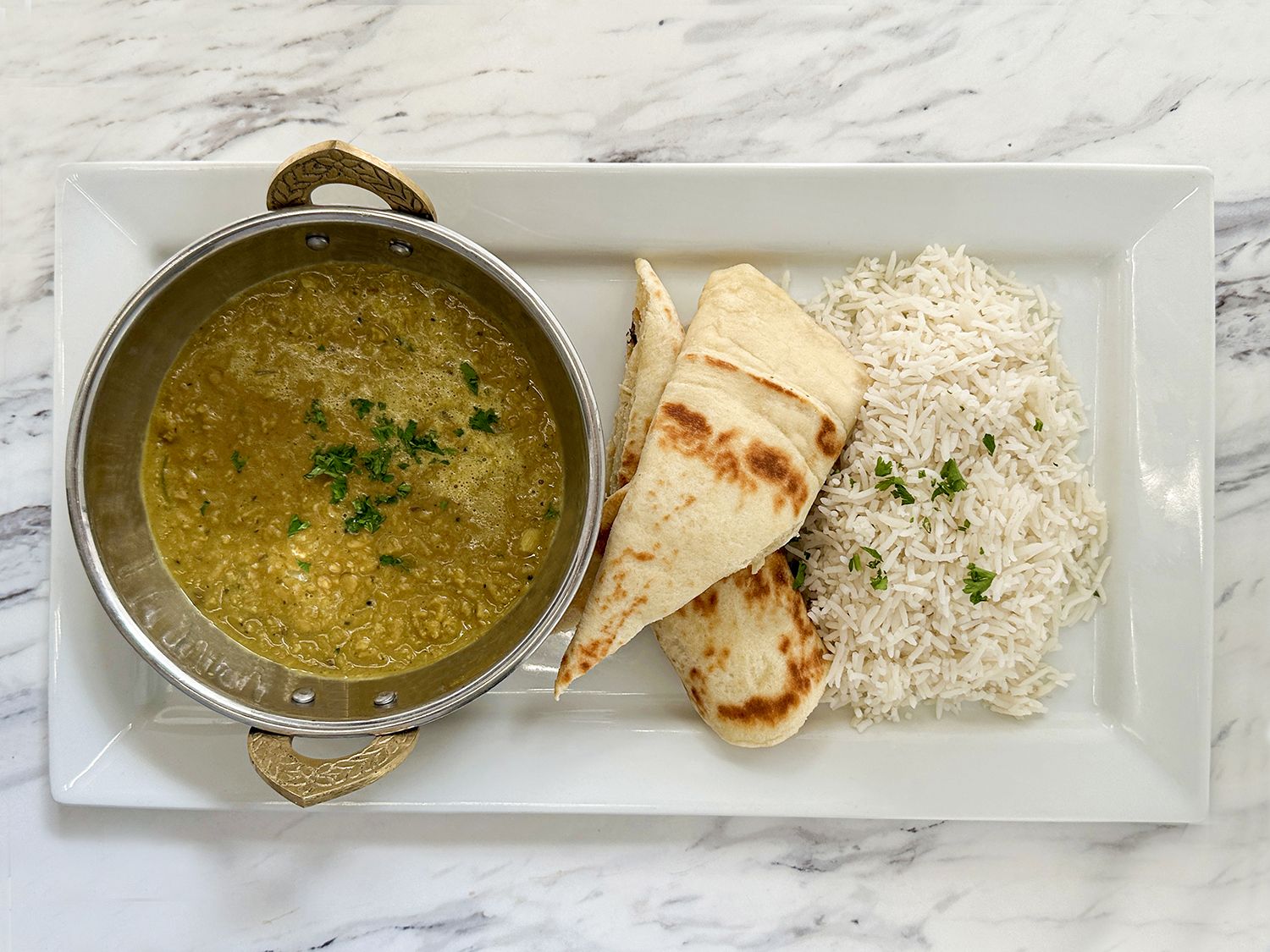 A metal bowl of green lentil curry with naan and white rice on a rectangular white plate over a marble surface.